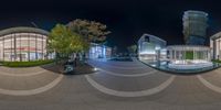 a street with a bench sitting next to a glass building at night, with the reflection of trees in the glass