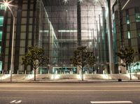 a glass building with several trees in the corner at night time near the street curb