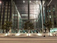 a glass building with several trees in the corner at night time near the street curb