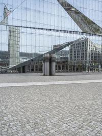 a building is reflected in the mirrored windows of a glass wall and stone pavement outside