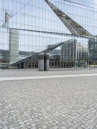 a building is reflected in the mirrored windows of a glass wall and stone pavement outside