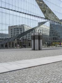 a building is reflected in the mirrored windows of a glass wall and stone pavement outside