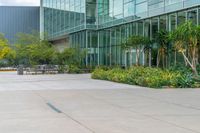 an empty courtyard that has benches and plants outside of it on a sunny day with a big glass building