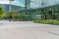 an empty courtyard that has benches and plants outside of it on a sunny day with a big glass building