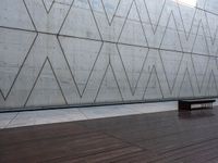 a wooden bench sits against a concrete wall that has geometric designs on it while others sit in the background