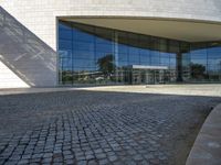 a modern building is featured behind a glass wall at the entrance of the building, which sits on a stone pathway