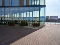 a clock sits outside a glassy building as people walk by in the background and the ground around the courtyard is cobblestone