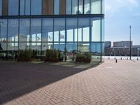 a clock sits outside a glassy building as people walk by in the background and the ground around the courtyard is cobblestone