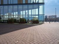 a clock sits outside a glassy building as people walk by in the background and the ground around the courtyard is cobblestone