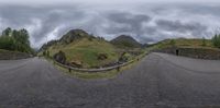 a road curves away from the camera under a gray sky with clouds above, and a stone bridge leading over to another area