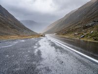 two sheep are standing by a curved road in the mountainside while cloudy skies gather overhead