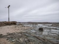 a street light with its base in the water beside rocks and sand shore line and an overcast sky