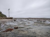 a wet beach with some rocks and water and a lighthouse in the distance under the cloudy sky