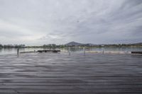 a wooden dock with benches under the water on the edge of a lake area with mountains and buildings on a cloudy day