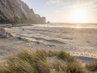 a beach with people sitting and walking near water at sunset under a mountain, rocks