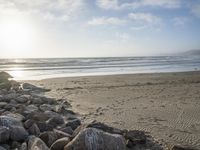 a surfer walks near rocks along the shore of a beach with a surfer in the distance