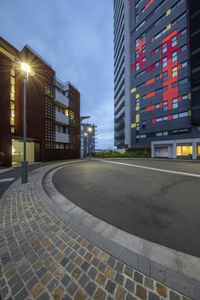 an empty street near some tall buildings in a city at night with brightly colored windows