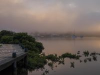 the dock sits on the water as the boats sit on the river side in the distance, and a beautiful pink cloud is in the sky