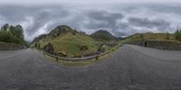 a road curves away from the camera under a gray sky with clouds above, and a stone bridge leading over to another area