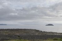 a body of water near a rocky shore and island in the distance as a cloudy sky above