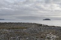 a body of water near a rocky shore and island in the distance as a cloudy sky above