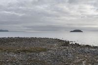 a body of water near a rocky shore and island in the distance as a cloudy sky above