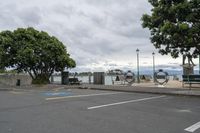 a street next to a parking space with benches and trees near it in front of the water