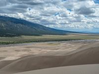 a sandy desert with mountains and a valley in the background under a cloudy blue sky