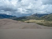 the view from behind of two sand dunes in front of mountains, clouds and a sky with low, overcast