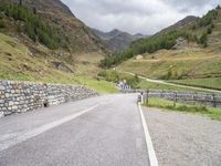 the road is going uphill in the mountains with a mountain pass by it and a stone bridge near the mountain