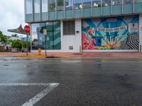 the wall on the building is covered with large murals and a yellow stop sign on a wet road