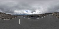 a curved road on the side of a mountain under cloudy skies and clouds with cars going underneath