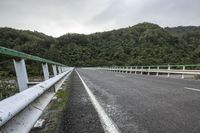 an empty road on a cloudy day in the countryside near trees and hills behind the bridge