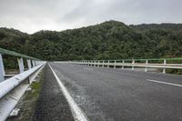 an empty road on a cloudy day in the countryside near trees and hills behind the bridge