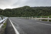 an empty road on a cloudy day in the countryside near trees and hills behind the bridge