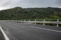 an empty road on a cloudy day in the countryside near trees and hills behind the bridge