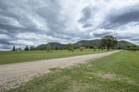 a dirt road that has gravel and grass and a mountain in the distance with clouds in the sky