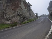 an empty road between large rocks in the foggy hillside with trees on each side