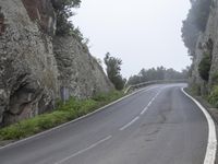 an empty road between large rocks in the foggy hillside with trees on each side