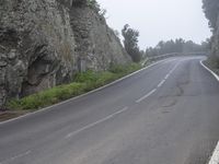 an empty road between large rocks in the foggy hillside with trees on each side