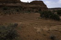 a dirt track running through a mountain with grass and trees along the sides, and a distant rocky landscape