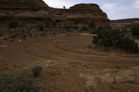 a dirt track running through a mountain with grass and trees along the sides, and a distant rocky landscape