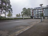 a empty, gray street in front of apartment buildings with trees, fencing and bushes