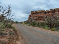 an empty dirt road lined with red rocks, trees, and bushes under a cloudy sky