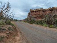 an empty dirt road lined with red rocks, trees, and bushes under a cloudy sky