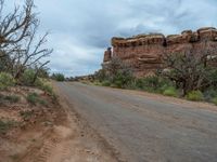 an empty dirt road lined with red rocks, trees, and bushes under a cloudy sky