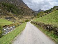 a gravel road running through a green valley with a stream in between two cliffs and grass on either side of the road