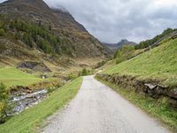 a gravel road running through a green valley with a stream in between two cliffs and grass on either side of the road