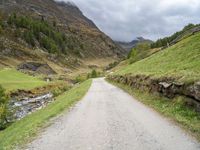 a gravel road running through a green valley with a stream in between two cliffs and grass on either side of the road