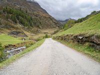 a gravel road running through a green valley with a stream in between two cliffs and grass on either side of the road